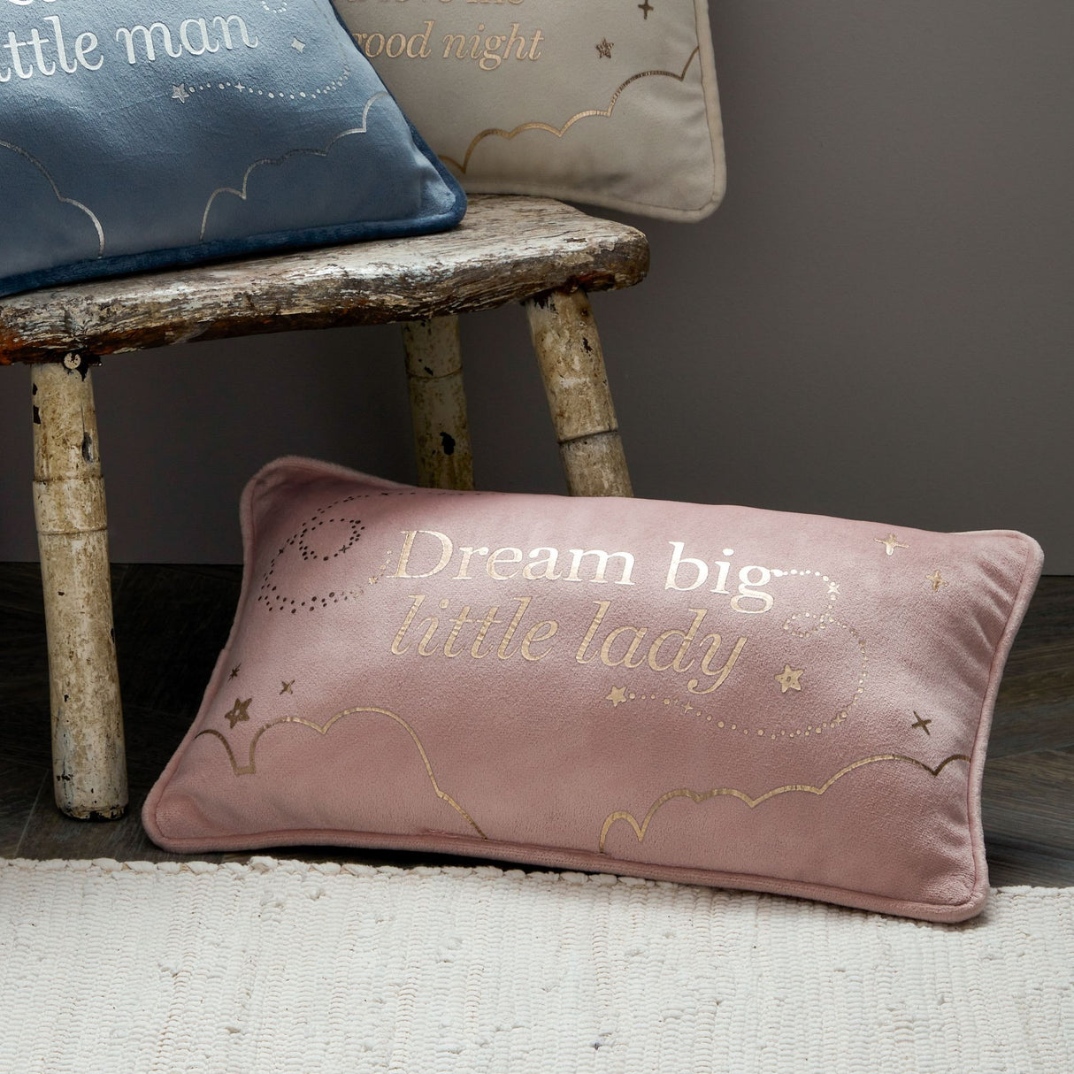 Dusty pink velvet rectangle cushion with "Dream big little lady" text and gold embroidered stars and clouds, displayed on rustic wooden bench with matching blue and cream pillows in neutral nursery setting