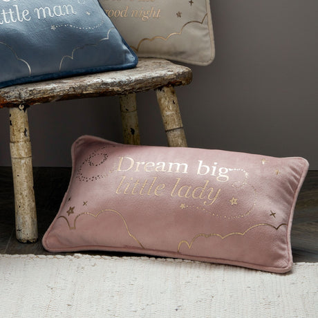 Dusty pink velvet rectangle cushion with "Dream big little lady" text and gold embroidered stars and clouds, displayed on rustic wooden bench with matching blue and cream pillows in neutral nursery setting