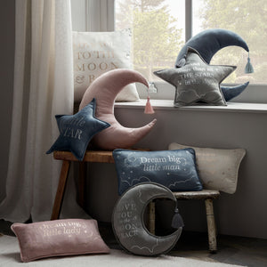 Pink velvet rectangular cushion with "Dream big little lady" text displayed among coordinating celestial-themed home decor including moon and star pillows on a wooden chair by a window