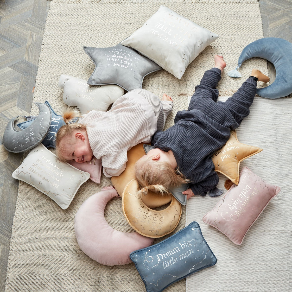 Two toddlers lying on a cream rug surrounded by various pastel-colored decorative pillows and cushions including pink crescent moon shape, blue star-shaped, and beige rectangular pillows with white embroidered text, demonstrating soft nursery bedding and baby room decor