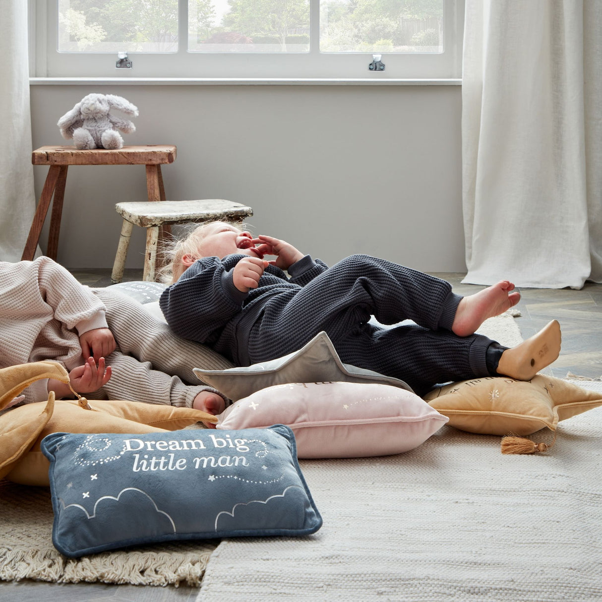 Two babies lying on soft cushions and blankets in a nursery room, with a blue velvet rectangle cushion reading "Dream big little man" in the foreground, wooden stools with a stuffed rabbit toy visible in the background near large windows