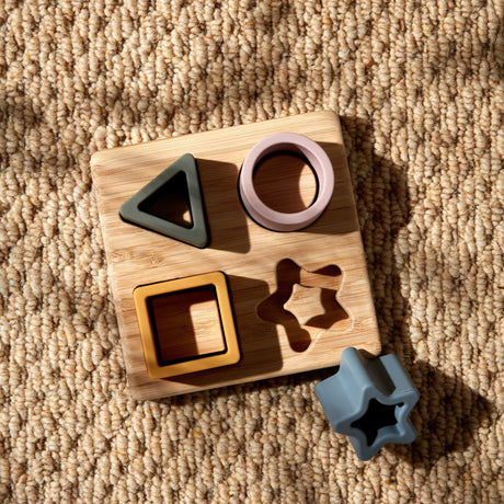 Bambino Silicone and Bamboo Star Puzzle featuring a natural bamboo board with four colorful silicone shape inserts in black, pink, yellow, and blue, displayed on a beige textured carpet with one blue star-shaped piece shown separately