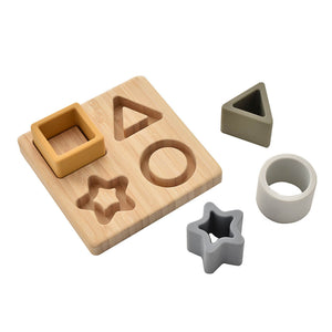 Bambino silicone and bamboo star puzzle toy featuring a natural bamboo board with carved shape cutouts and five removable silicone pieces in mustard yellow, grey, and white including square, triangle, circle, star and cylindrical shapes