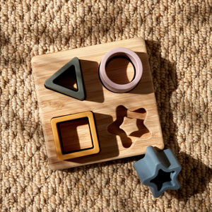 Bambino silicone and bamboo star puzzle featuring a natural wooden board with four colorful rubber shape cutouts in black, pink, yellow, and gray, displayed on a beige textured carpet