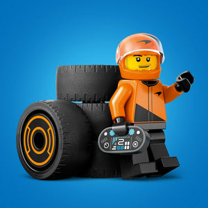 LEGO minifigure racing driver in bright orange helmet and suit with black gloves, standing next to large black racing tires with orange trim and a detailed gray digital dashboard, against a blue background