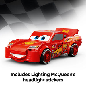 Red LEGO Lightning McQueen car model #77255 with blue eyes, yellow racing details, and black wheels, featuring an openable cargo bed, displayed against a grey checkered background with text indicating included headlight stickers