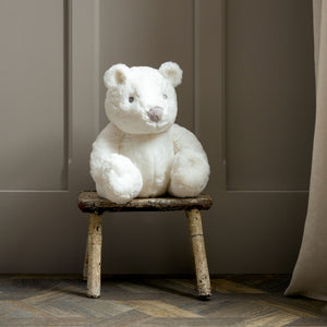 Large white plush teddy bear sitting on rustic wooden stool against neutral beige wall backdrop