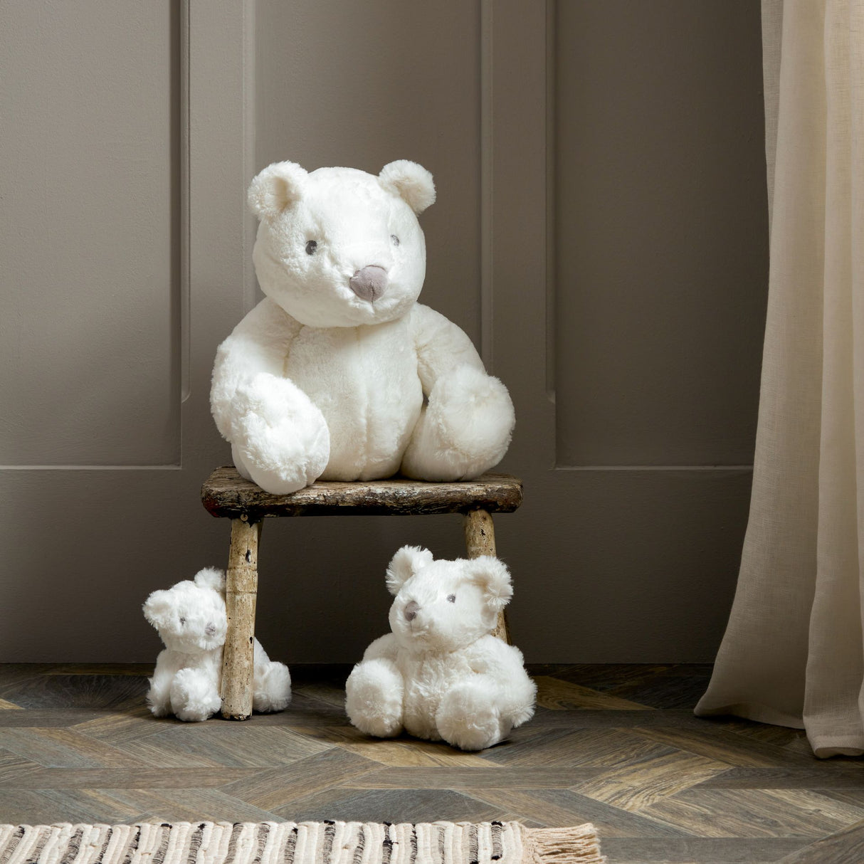 Large white plush teddy bear sitting on rustic wooden bench with two smaller white teddy bears on floor beside it in modern minimalist interior setting with neutral beige and grey tones