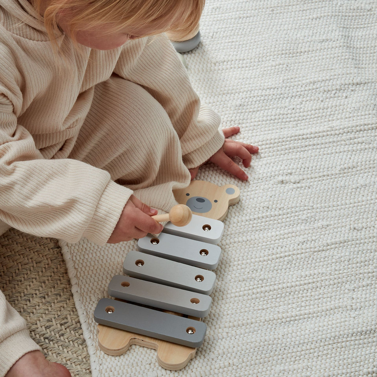 Baby playing with wooden teddy bear xylophone toy featuring gray metal bars and natural wood base on white textured blanket