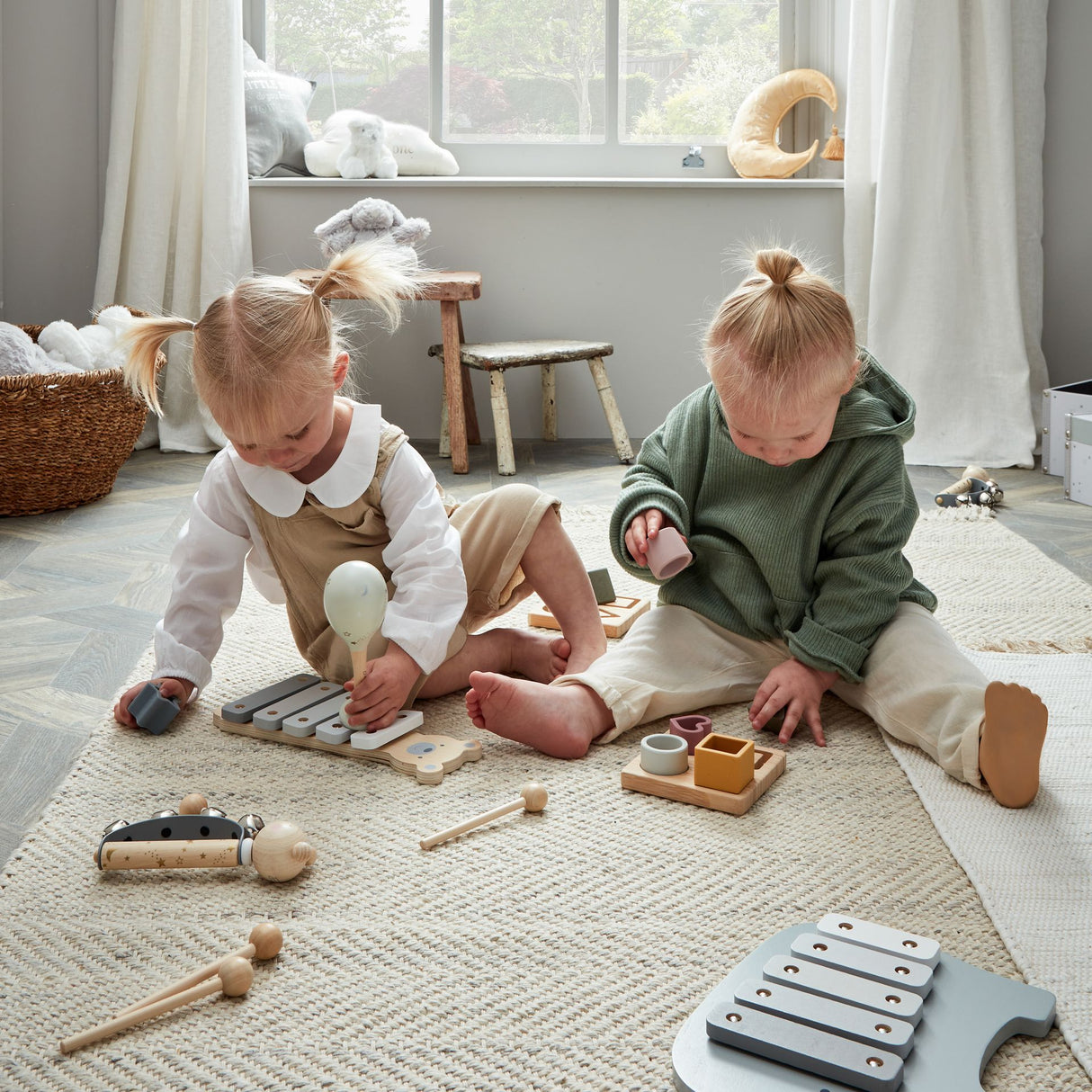 Two toddlers playing with wooden musical instruments including a grey xylophone in a bright, minimalist Scandinavian-style nursery with natural wooden toys, wicker baskets, and large windows overlooking greenery