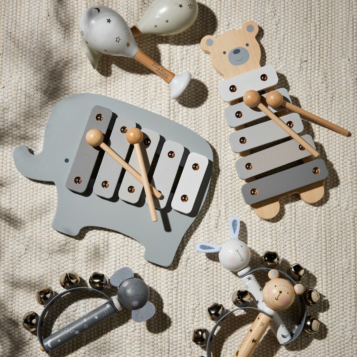Bambino wooden toy xylophone set displayed on cream fabric including grey elephant-shaped xylophone with wooden mallets, bear-shaped xylophone, star-patterned maracas, wooden rattles with bells and natural wood beads in grey, white and natural timber finishes