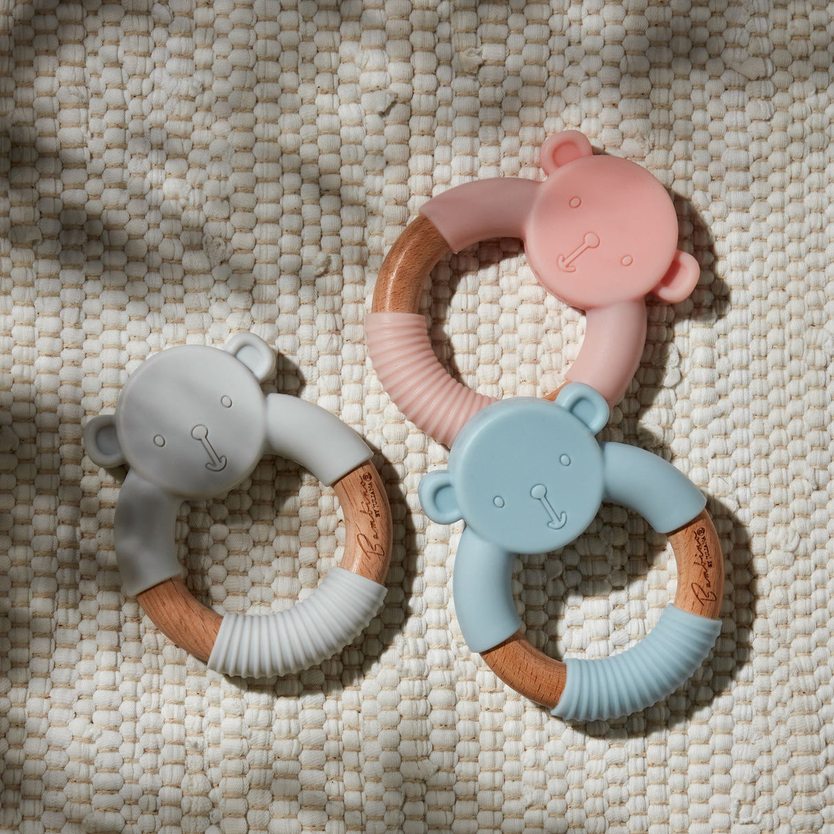 Three soft silicone teething rings with wooden handles in gray, pink, and blue, each featuring a cute teddy bear head design with embossed facial features, arranged on a cream textured fabric background