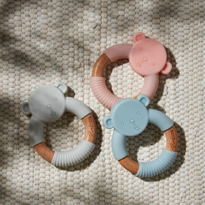 Three soft silicone teething rings with wooden handles in gray, pink, and blue, each featuring a cute teddy bear head design with embossed facial features, arranged on a cream textured fabric background
