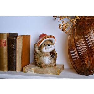 Wooden hedgehog figurine wearing a red and white Santa hat, displayed on a cream-colored book between antique leather-bound books and a brown woven vase with dried flowers on a white shelf