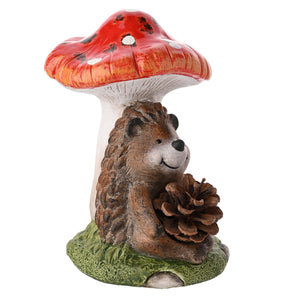 Decorative garden figurine of a brown hedgehog holding a pinecone beneath a red and white toadstool mushroom on a green grass base, whimsical country style ornament