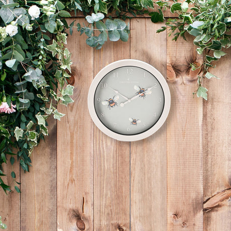 Country Living outdoor clock with bee design featuring white frame and grey face with illustrated bees instead of numbers, mounted on wooden garden wall with climbing ivy and white flowers