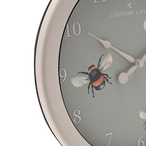 Close-up view of a Country Living outdoor clock with a grey dial featuring a colorful bee illustration, white numbers 6-10 visible, white metal hands, cream-colored rim, and black bezel, 26.5cm diameter
