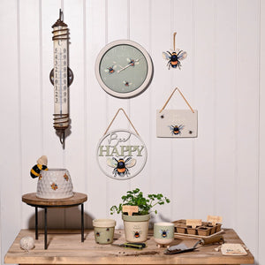 Country Living bee-themed outdoor clock display featuring a 26.5cm round sage green clock with bee motifs, surrounded by coordinating bee décor including a thermometer, hanging bee ornaments, ceramic planters, candles, and a decorative beehive cake on a wooden table against white shiplap wall