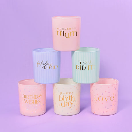 Collection of six pastel-colored scented candles arranged in a pyramid formation against a purple background, featuring gold text on pink, blue, mint green and cream containers with messages including "Wonderful Mum," "Fabulous Friend," "You Did It," "Birthday Wishes," "Happy Birthday," and "Love"
