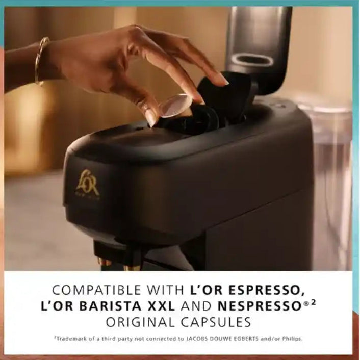 Hand inserting a coffee capsule into a black Philips L'Or Barista coffee machine with gold accents, shown in a modern kitchen setting with compatibility text for L'Or Espresso, L'Or Barista XXL and Nespresso Original capsules