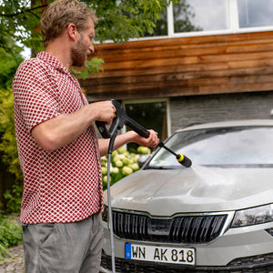 Man washing a silver Skoda car with a pressure washer in front of a house.