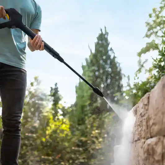 Person using Karcher K7 WCM Flex Eco Booster pressure washer to clean brick wall with water spray, surrounded by green trees and foliage on sunny day