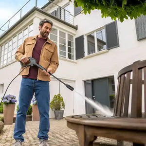 Man in tan jacket and blue jeans using Karcher K3 pressure washer with lance and hose to clean patio outside modern white house with potted plants
