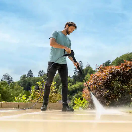Man in light blue t-shirt and dark pants using a Karcher K7 pressure washer with booster to clean a concrete patio, with evergreen trees and garden in background under blue sky