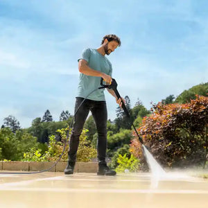 Man in light blue t-shirt and dark pants using a Karcher K7 pressure washer with booster to clean a concrete patio, with evergreen trees and garden in background under blue sky