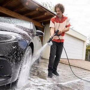 Man in red and cream shirt using Karcher K3 pressure washer with blue hose to clean black car in driveway, with garage in background