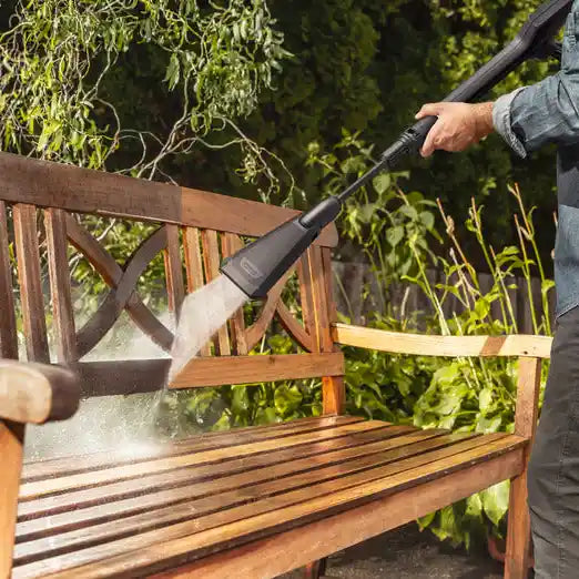 Man using Karcher K7 WCM Flex Eco Booster pressure washer to clean wooden garden bench with high-pressure water spray, with green foliage visible in background