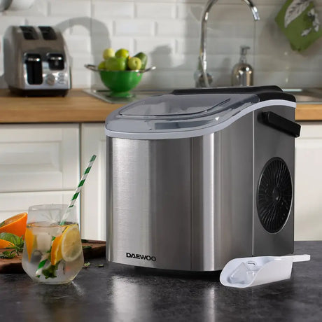 Daewoo stainless steel ice cube maker with 1.2 litre tank on kitchen counter next to glass of iced lemonade with orange slices, displaying silver and black design with control panel and scoop