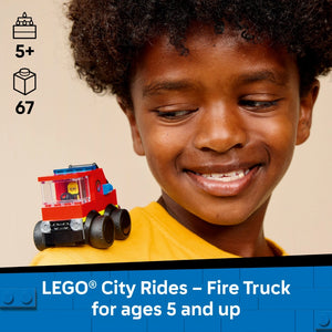 A smiling child wearing a yellow shirt holds a red LEGO City fire truck with black wheels and blue details, displayed against a beige background with age rating 5+ and 67 piece count indicators