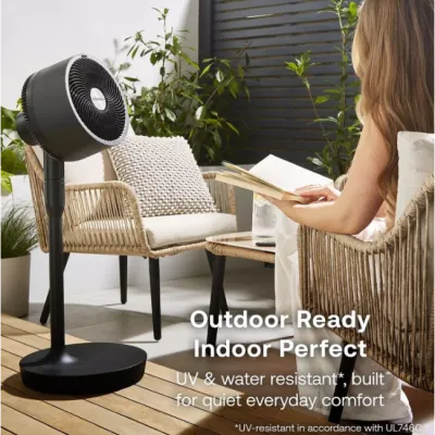 Black Dimplex Duocool 2-in-1 pedestal fan on outdoor patio with woman in white top sitting in woven rattan chair, surrounded by potted plants, with text overlay stating Outdoor Ready Indoor Perfect UV and water resistant