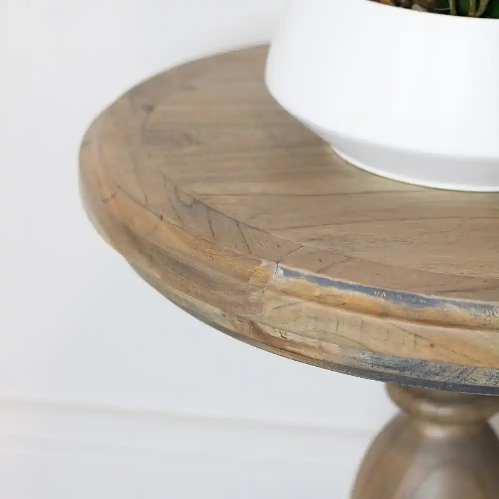 Close-up detail of a round wooden side table with natural light wood finish, showing carved edge detail and metal banding, topped with a white ceramic planter containing green foliage