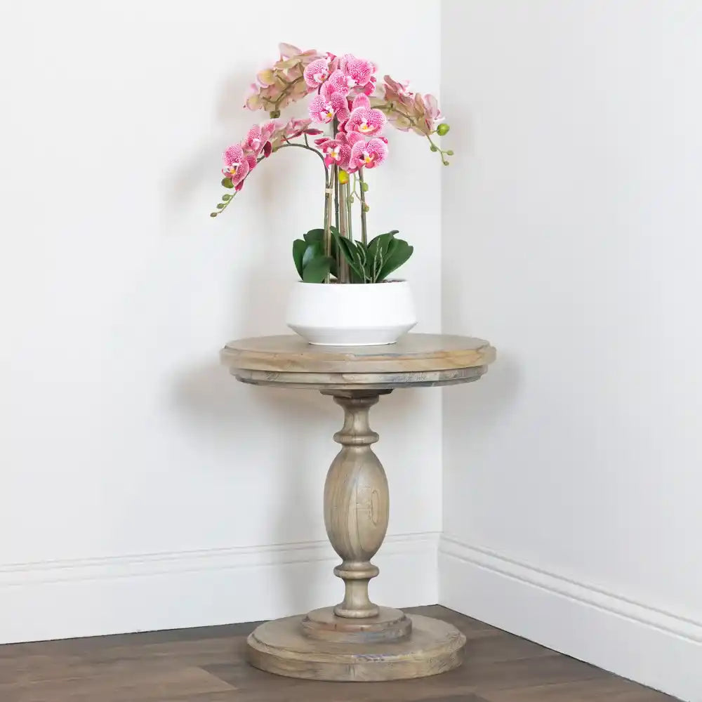 Tara Lane round pillar side table in weathered wood with pink orchid arrangement in white pot, positioned in corner against white walls and dark wood flooring