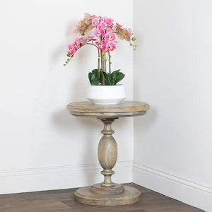 Tara Lane round pillar side table in weathered wood with pink orchid arrangement in white pot, positioned in corner against white walls and dark wood flooring