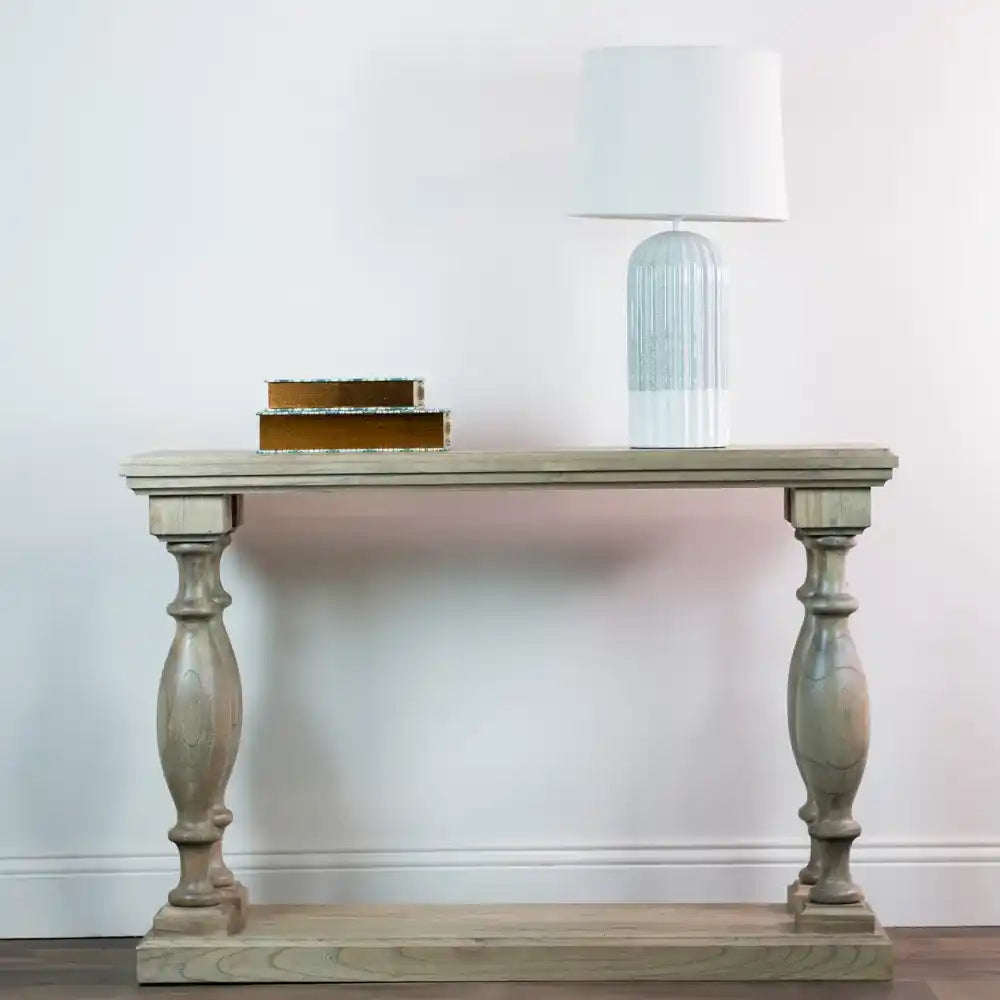 Tara Lane Pillars Console Table in whitewashed wood with ornate turned legs, styled with stacked gold-edged books and a blue glass table lamp with white shade against a white wall