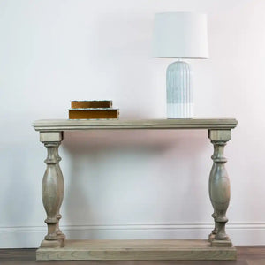 Tara Lane Pillars Console Table in whitewashed wood with ornate turned legs, styled with stacked gold-edged books and a blue glass table lamp with white shade against a white wall