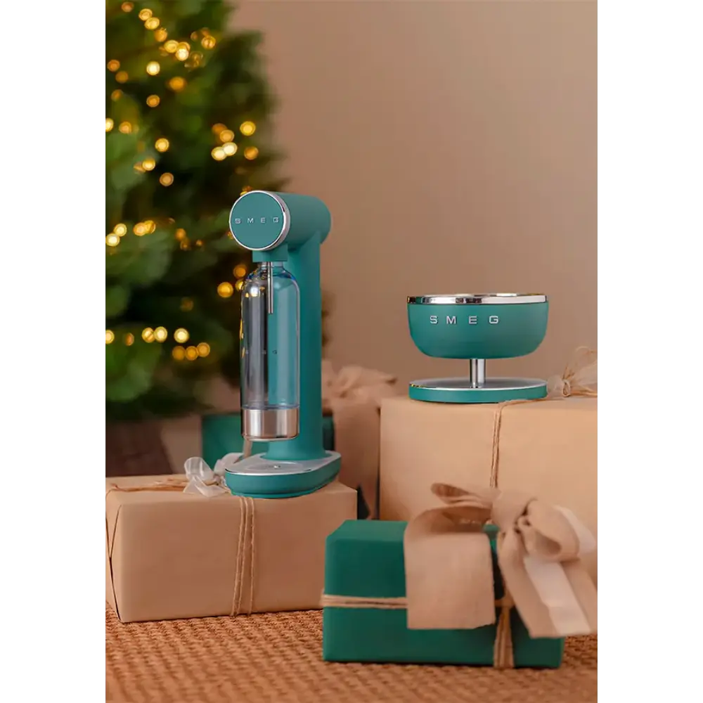 Smeg emerald green sparkling water soda maker with glass bottle displayed on kraft gift boxes next to a matching bowl, styled with Christmas decorations and twinkling lights in the background