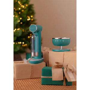 Smeg emerald green sparkling water soda maker with glass bottle displayed on kraft gift boxes next to a matching bowl, styled with Christmas decorations and twinkling lights in the background
