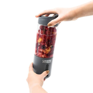 Urban Fitness VitalFuse portable blender in grey being held by two hands, containing a red smoothie with fruit pieces inside, product shot against white background