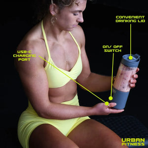 Athletic woman in yellow sports bikini holding grey portable blender with blue lid, demonstrating USB-C charging port and on/off switch against black background, Urban Fitness product showcase