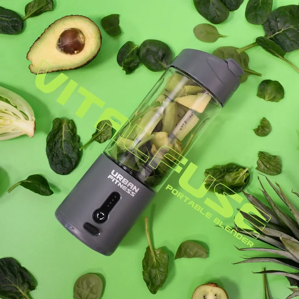 Urban Fitness VitalFuse portable blender in grey with black accents surrounded by fresh green vegetables and herbs including avocado, spinach, lettuce and spring onions on a bright green background