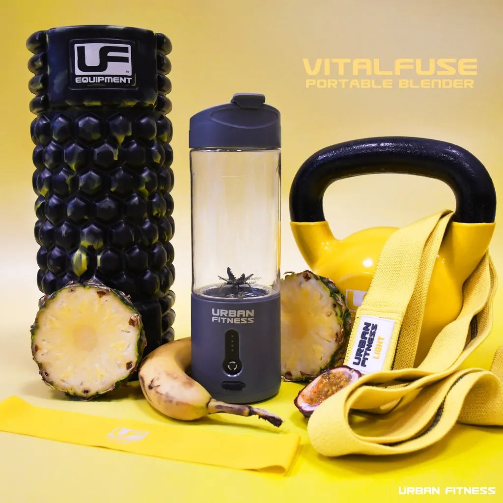 Urban Fitness VitalFuse portable blender in dark navy blue and black with clear glass pitcher, displayed on yellow background alongside fresh pineapple, banana, yellow resistance band and fitness accessories