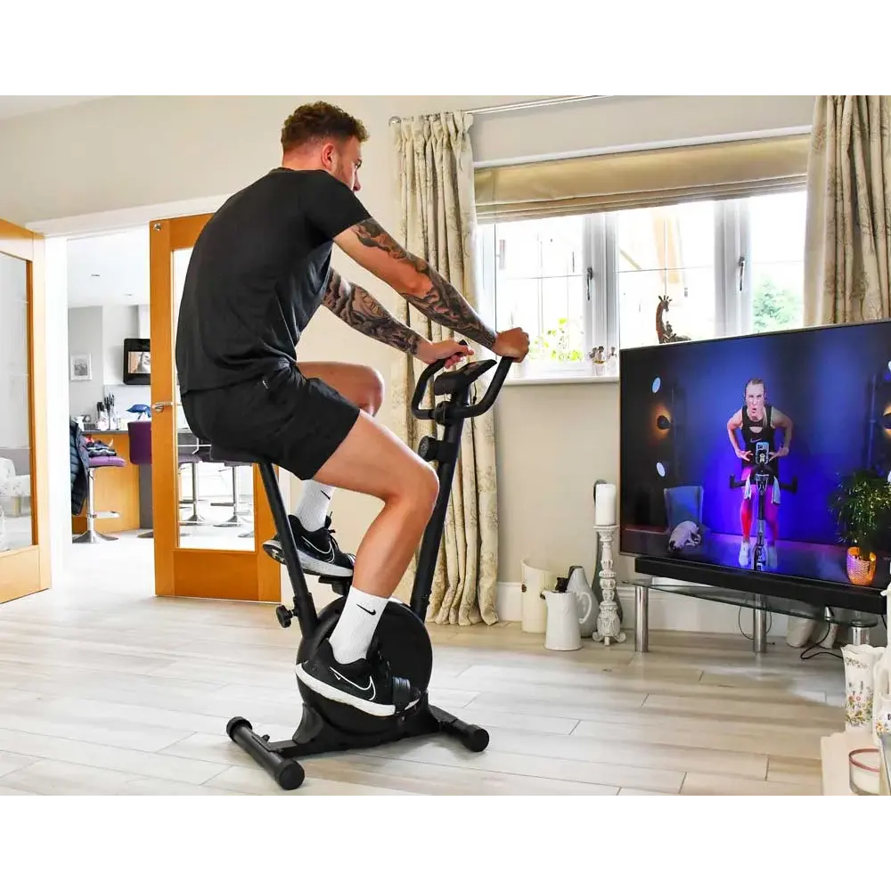Man with tattoos exercising on black Urban Fitness magnetic stationary bike in modern living room while watching fitness instructor on TV screen