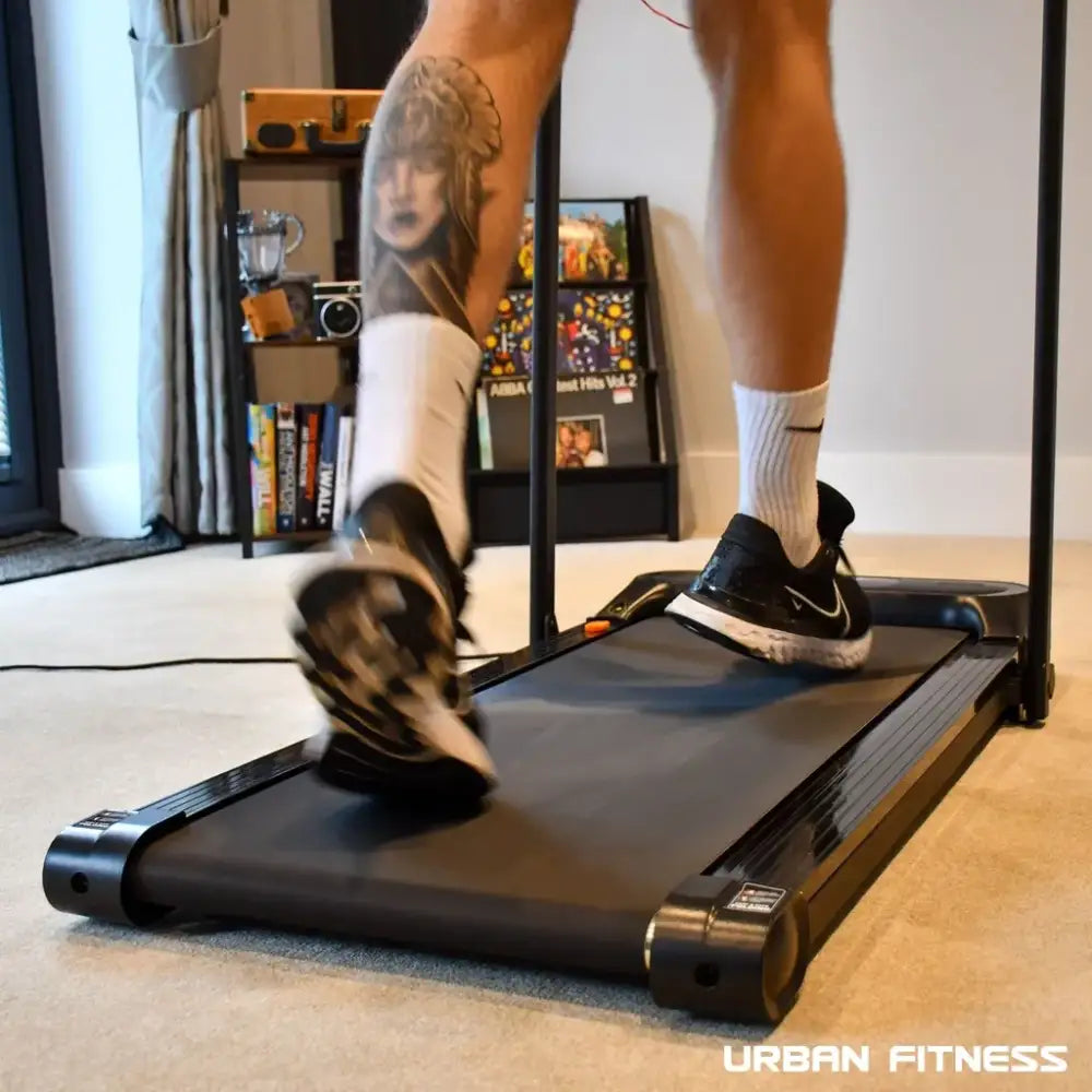 Person running on a black Urban Fitness walking treadmill in a modern home gym with white socks and black Nike shoes, shelving with decorative items visible in the background