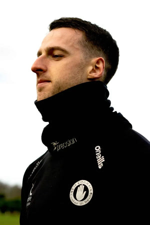 Man wearing black Precision Essential Neck Warmer Snood with embroidered logo and branding, shown in profile against white background in outdoor setting