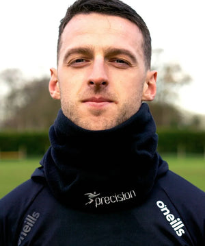 Man wearing black Precision Essential Neck Warmer Snood with Precision and O'Neills branding, outdoor setting with green grass and blurred background
