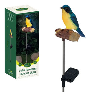 Gardenkraft Solar Tweeting Bluebird Light showing product packaging on left and assembled garden stake light on right featuring a blue and yellow bird figurine perched on a brown textured branch with black solar panel base